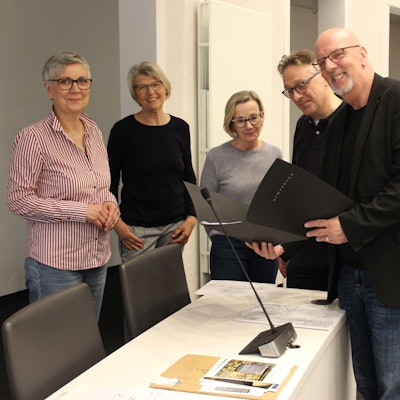 Männer und Frauen stehen an einem Tisch und halten schwarze Mappen.