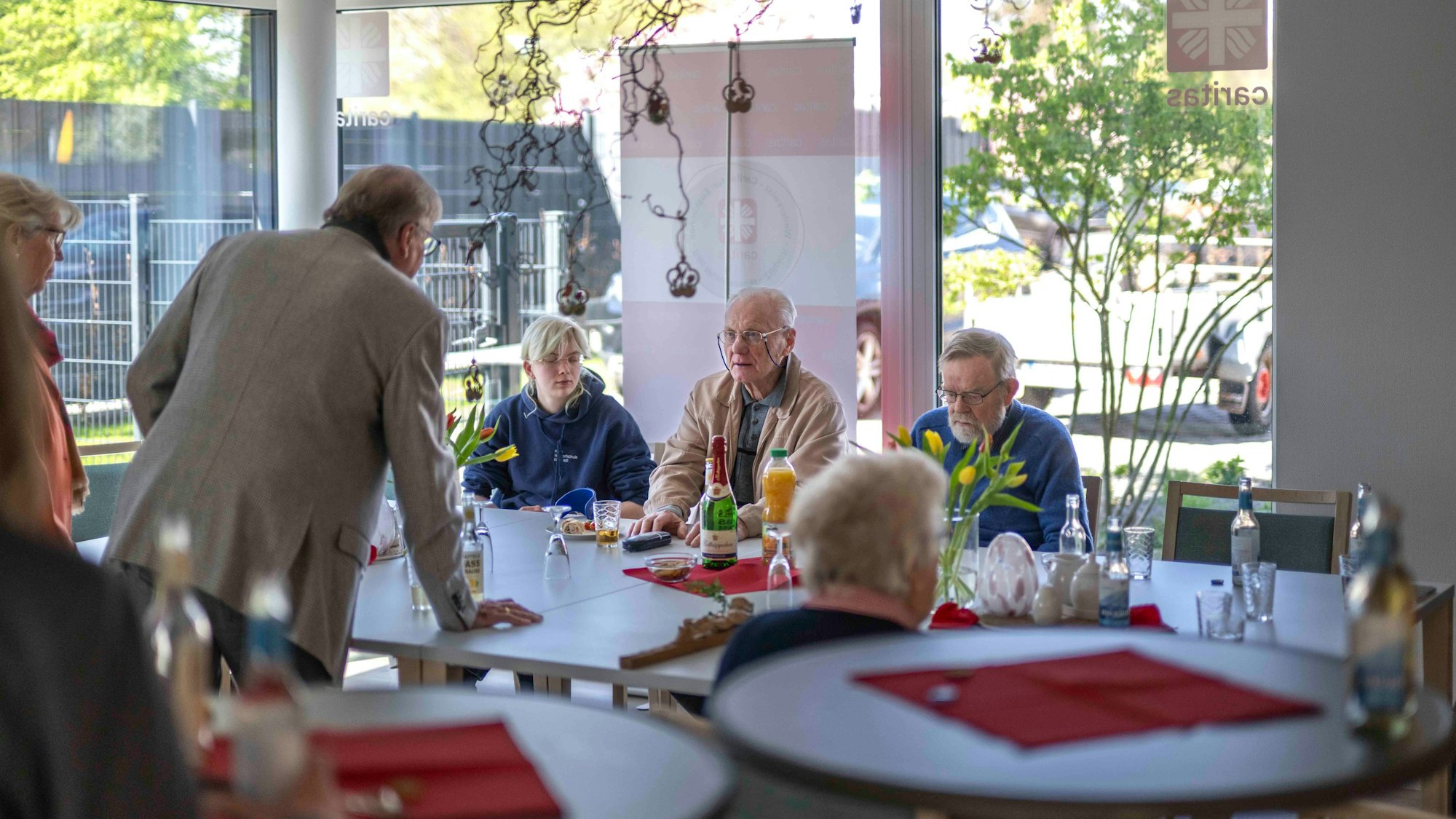 Blick in die lichthelle Tagesstätte. Ein paar Gäste sitzen am Tisch, der mit Blumen geschmückt ist.