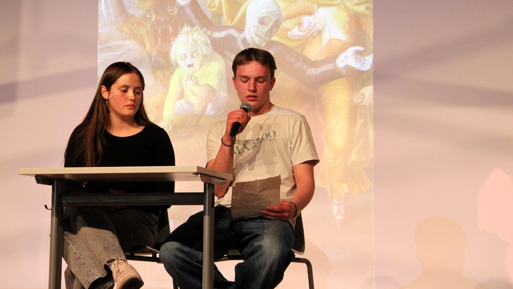 Ein Mädchen und ein Junge sitzen an einem Tisch, der junge Mann liest etwas vor.