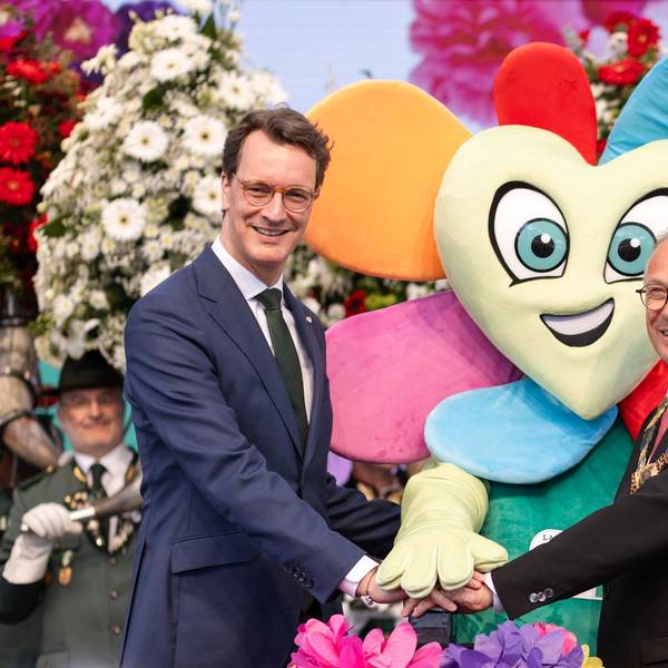 Zwei Männer drücken auf einen Buzzer. Hinter ihnen steht ein Maskottchen in Form einer Blume.
