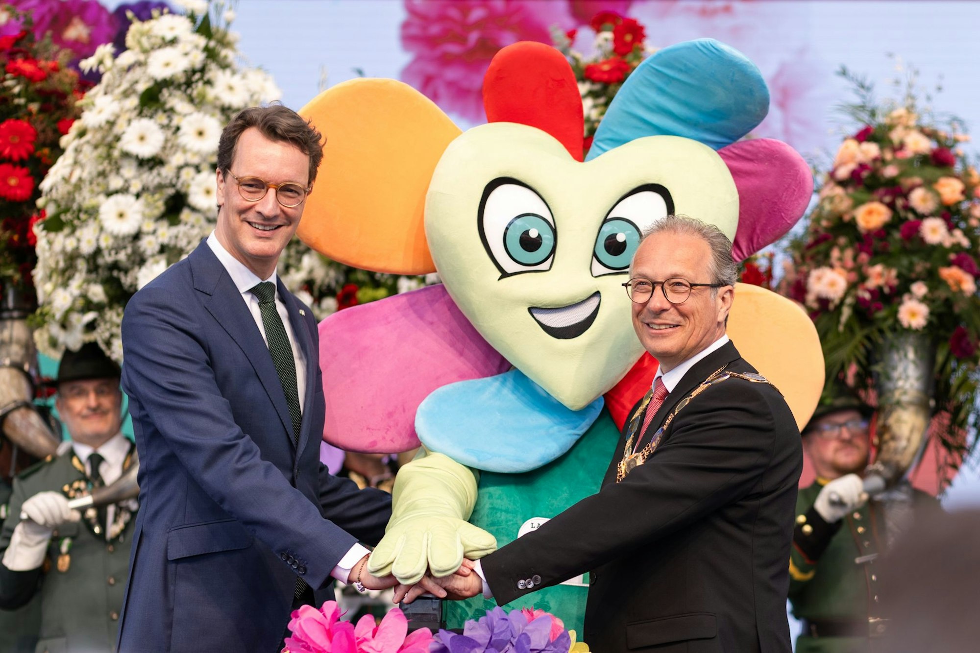 Zwei Männer drücken auf einen Buzzer. Hinter ihnen steht ein Maskottchen in Form einer Blume.