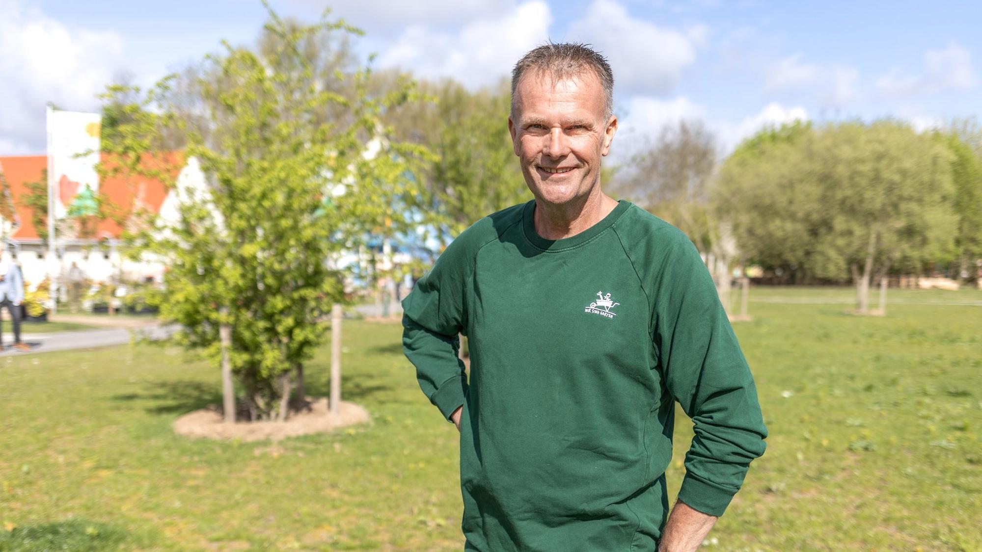 Ein Mann im grünen Pullover steht auf einer Wiese und schaut in die Kamera.