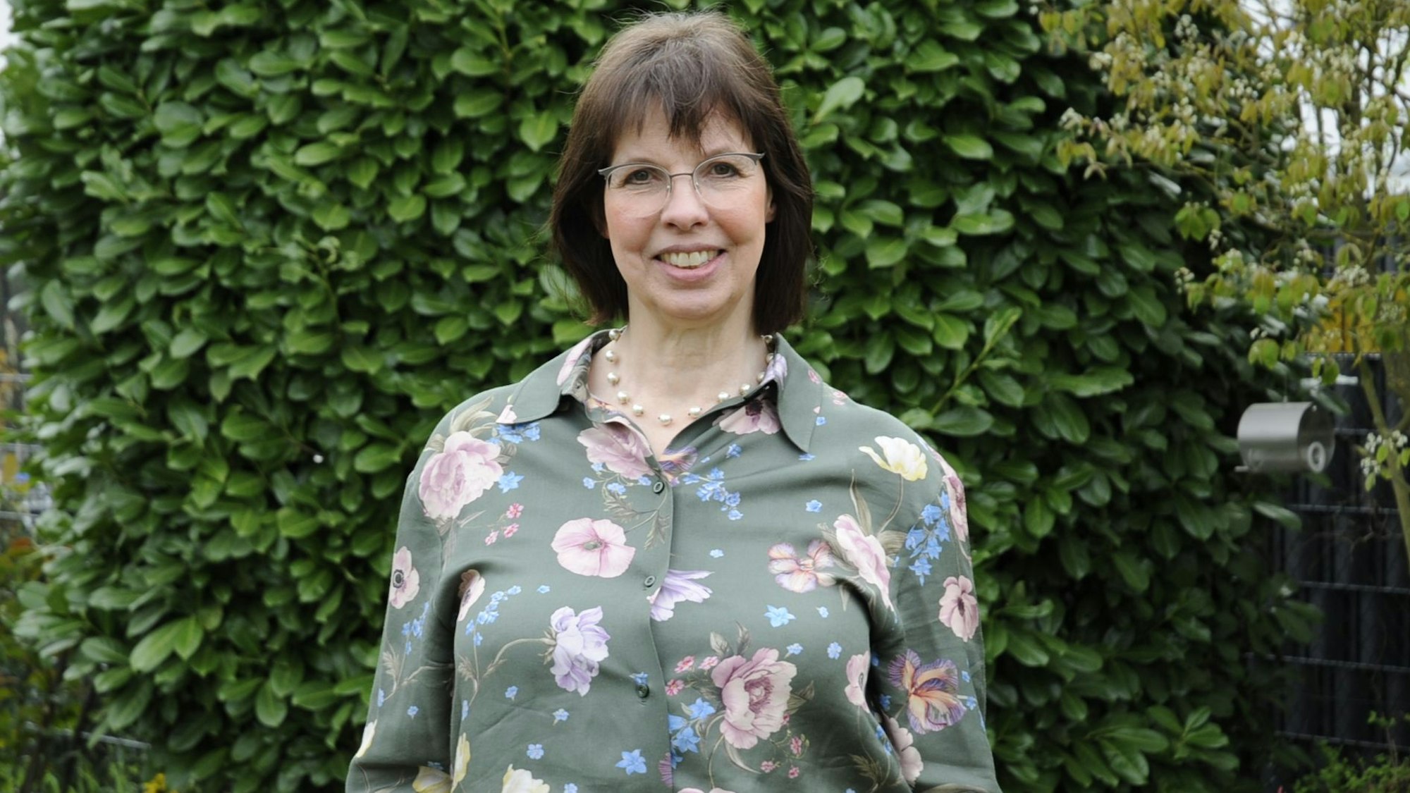 Eine Frau mit dunklen Haaren steht vor einer Hecke.