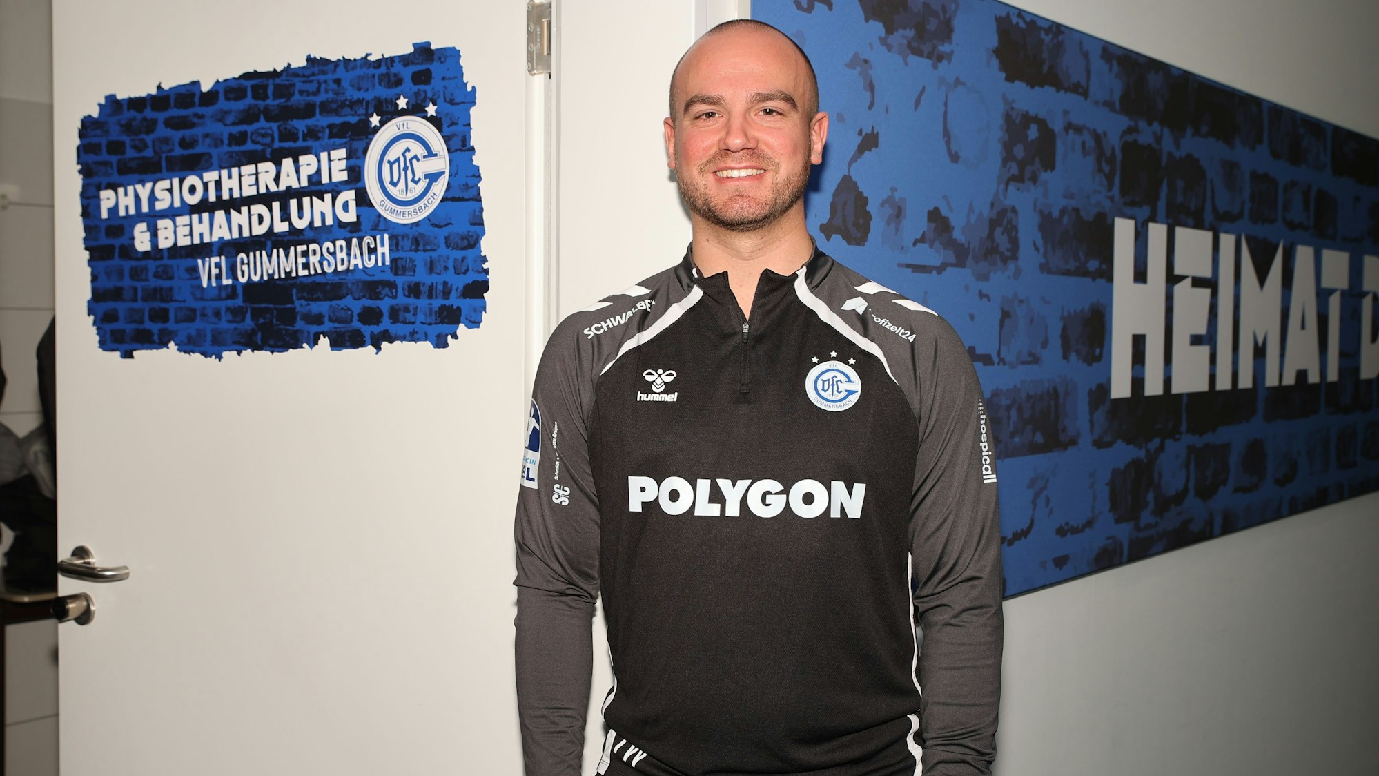 Ein Mann steht vor einer Kabine, anderen Tür die Aufschrift „Physiotherapie“ zu lesen ist.