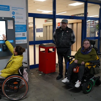 Zu sehen sind zwei Frauen im Rollstuhl sowie ein Mann, sie befinden sich an einem Parkautomaten.