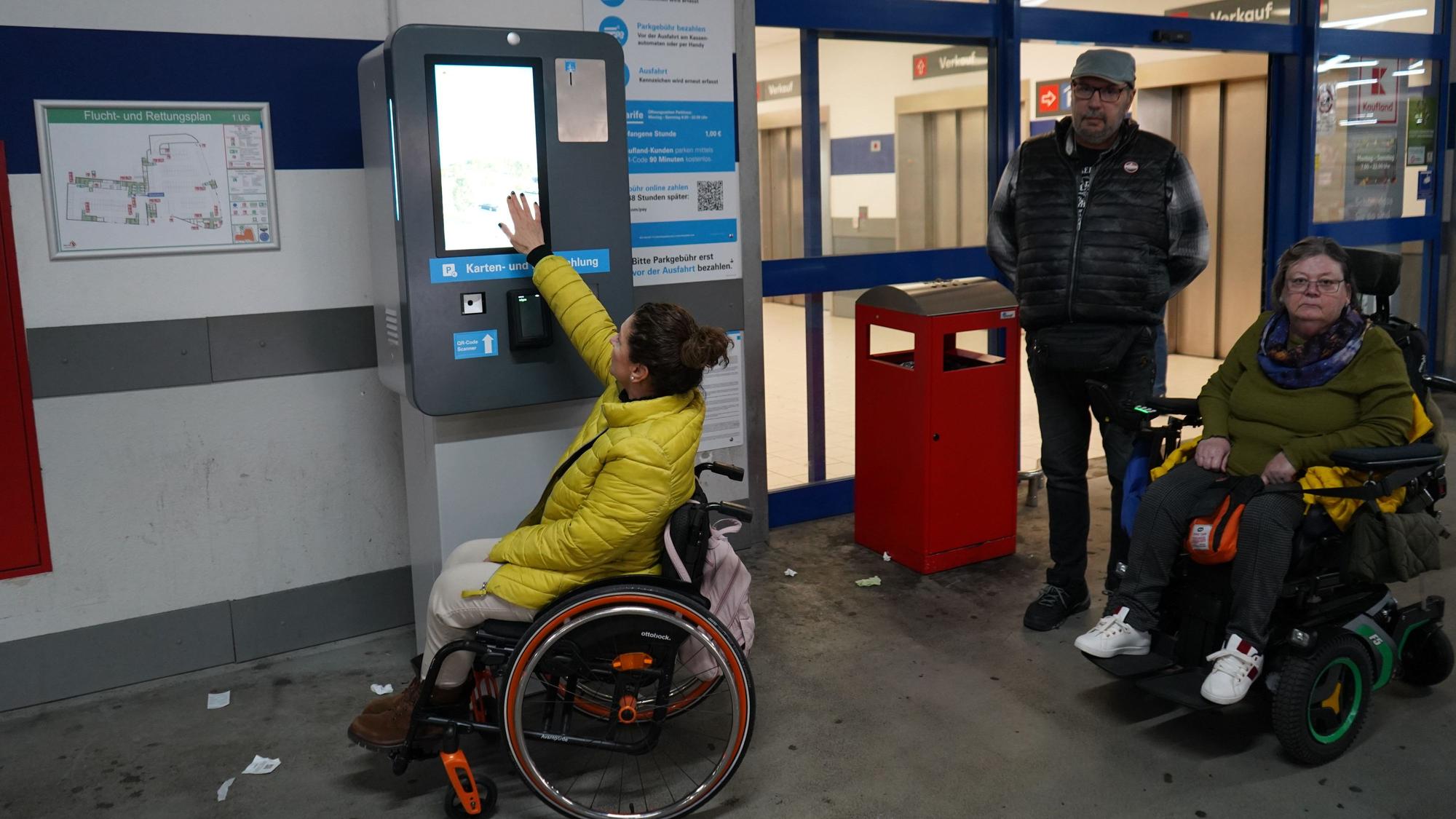 Zu sehen sind zwei Frauen im Rollstuhl sowie ein Mann, sie befinden sich an einem Parkautomaten.