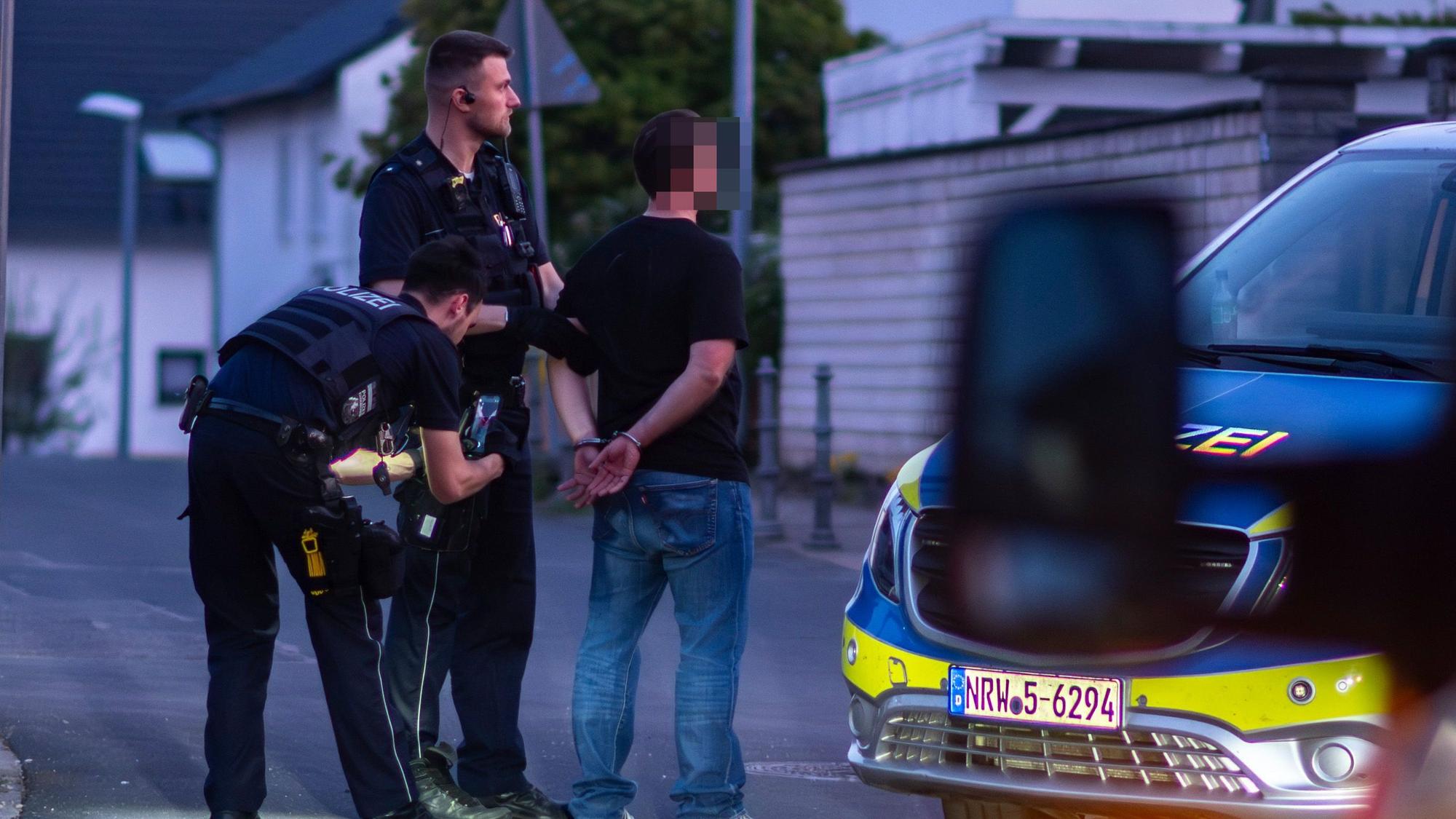 Das Bild zeigt einen Mann, der seine Hände hinter dem Rücken mit Handschellen gefesselt hat. Auch zwei Polizisten sind zu sehen.