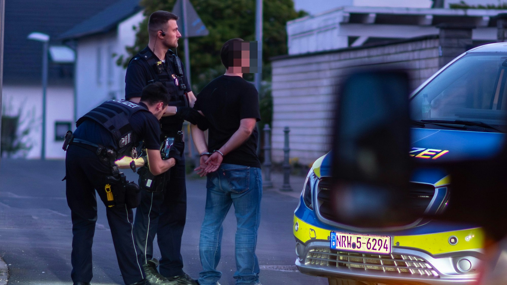 Das Bild zeigt einen Mann, der seine Hände hinter dem Rücken mit Handschellen gefesselt hat. Auch zwei Polizisten sind zu sehen.