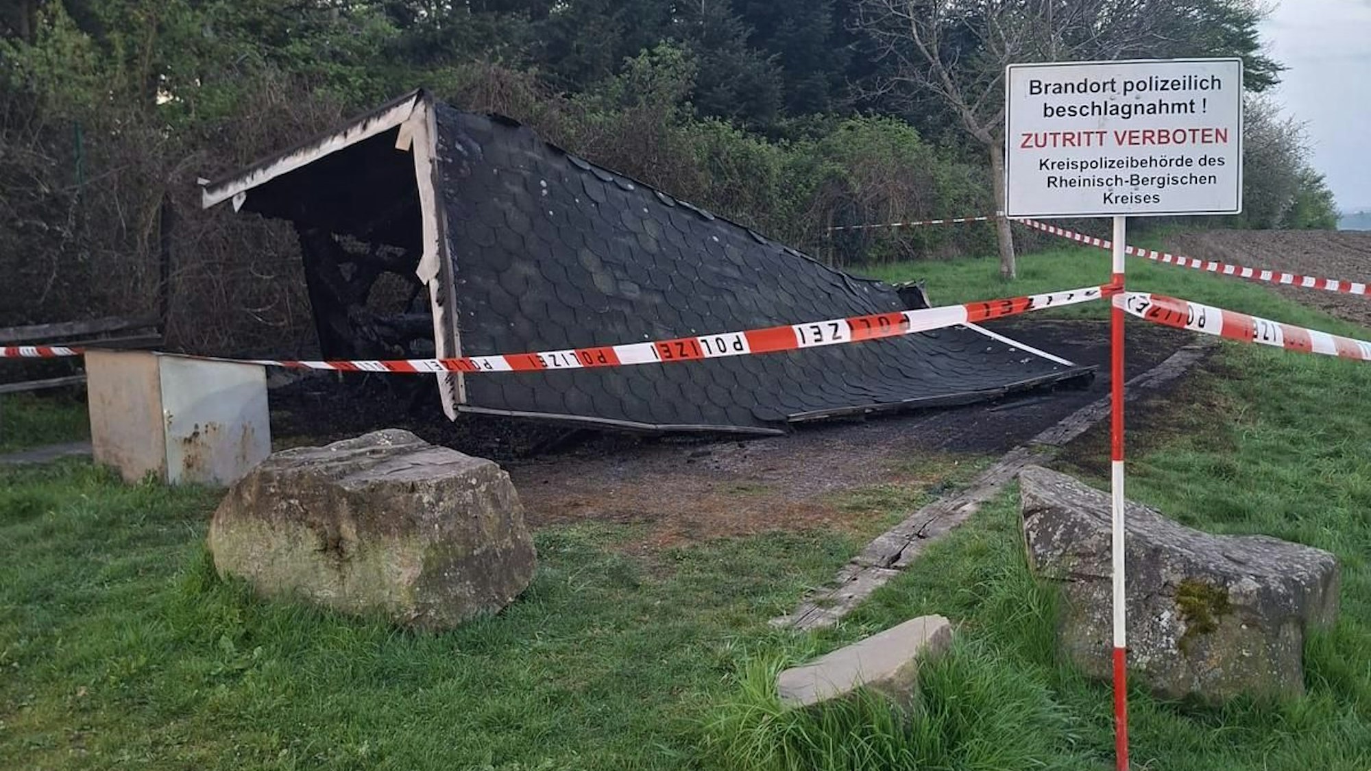 Eine Polizeiabsperrung steht vor einer abgebrannten Holzhütte.