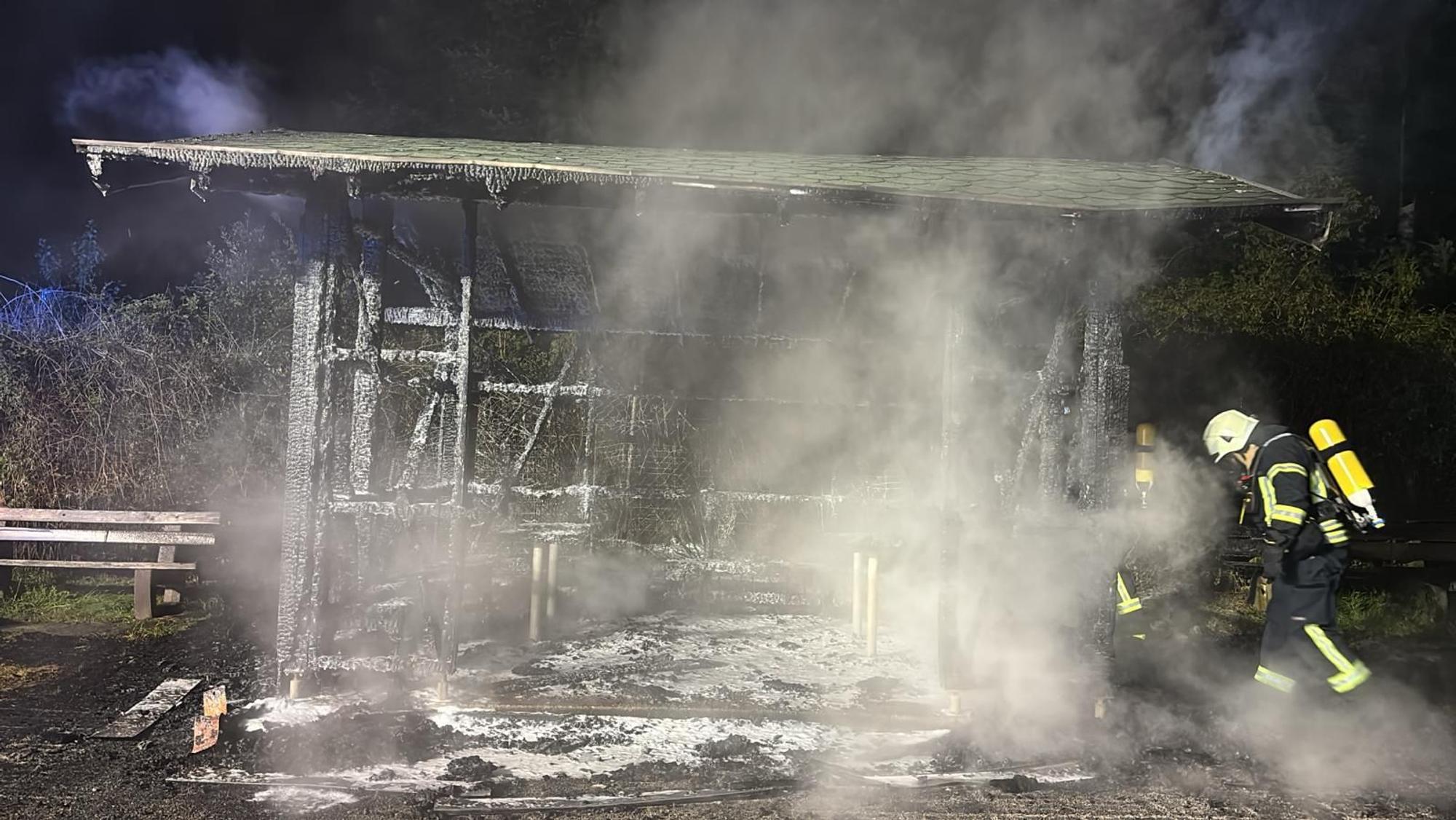 ein Feuerwehrmann steht neben den rauchenden Trümmern der Wanderhütte bei Overath-Eulenthal.