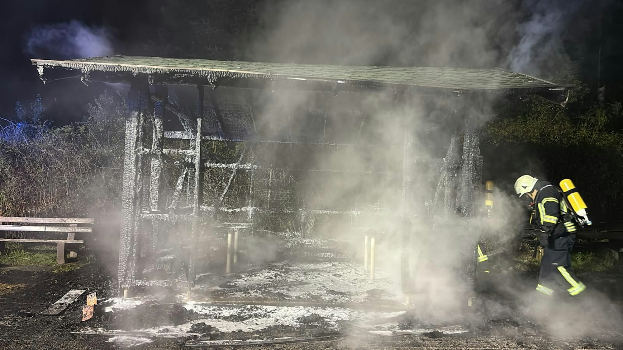 ein Feuerwehrmann steht neben den rauchenden Trümmern der Wanderhütte bei Overath-Eulenthal.