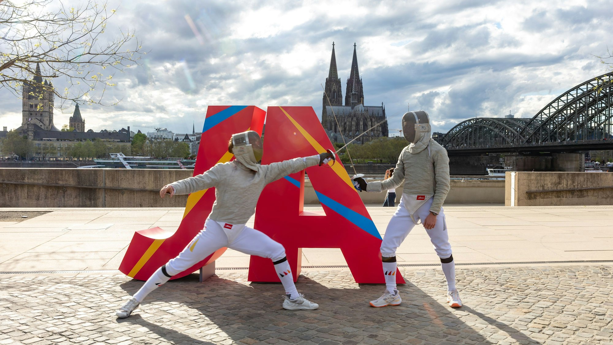 Zwei Fechter stehen vor einem „Ja“-Schriftzug am Rheinufer.