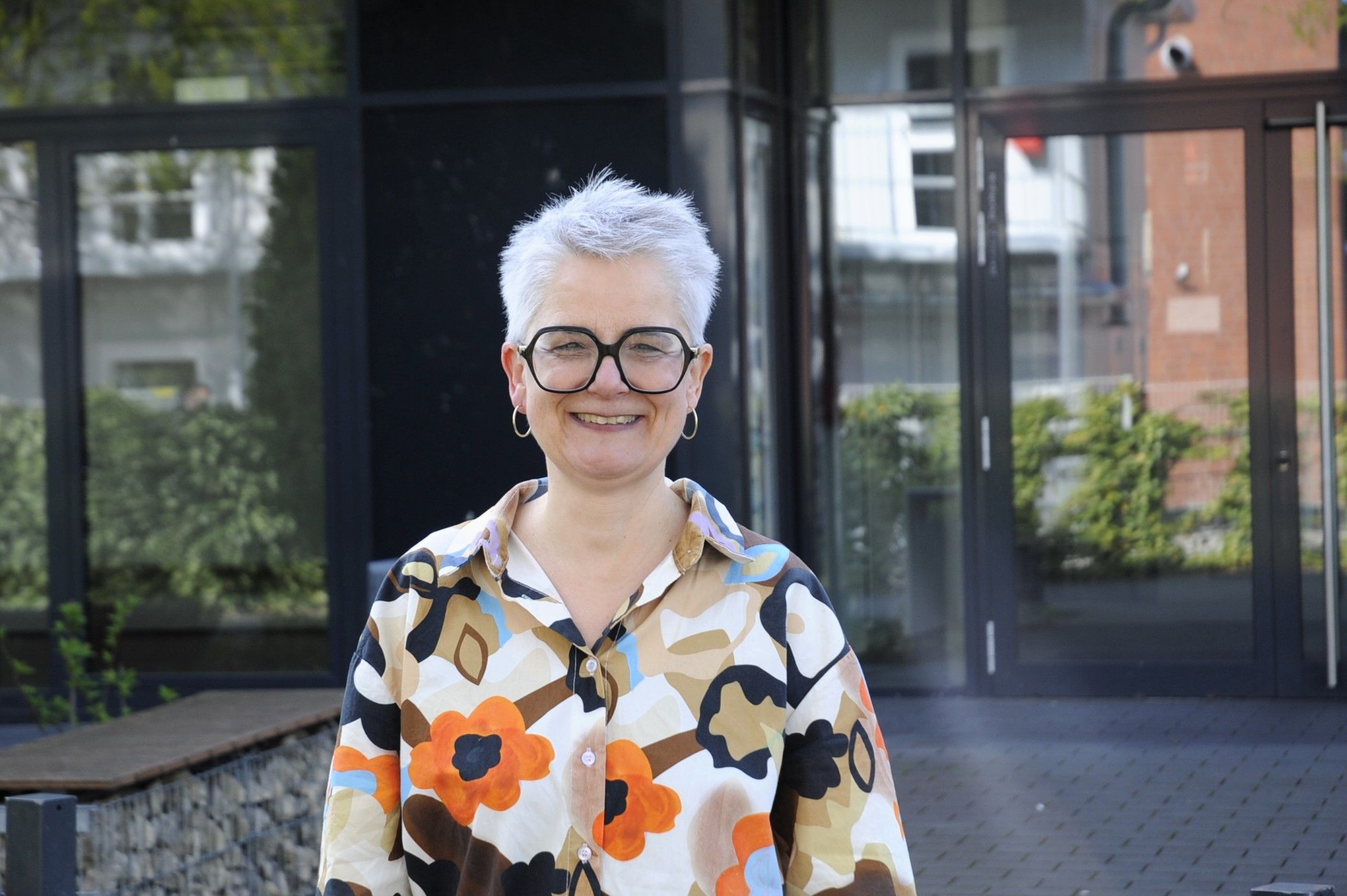 Eine Frau mit Brille und kurzen Haaren steht vor einem Gebäude.