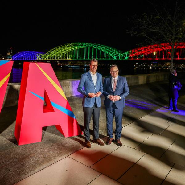 Hendrik Wüst (CDU, l.), NRW-Ministerpräsident, und Torsten Burmester (SPD), Oberbürgermeister der Stadt Köln, stehen vor dem großen „Ja“ und der in den olympischen Farben angeleuchteten Hohenzollernbrücke.