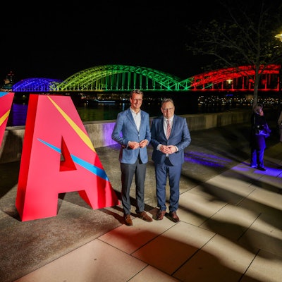 Hendrik Wüst (CDU, l.), NRW-Ministerpräsident, und Torsten Burmester (SPD), Oberbürgermeister der Stadt Köln, stehen vor dem großen „Ja“ und der in den olympischen Farben angeleuchteten Hohenzollernbrücke.