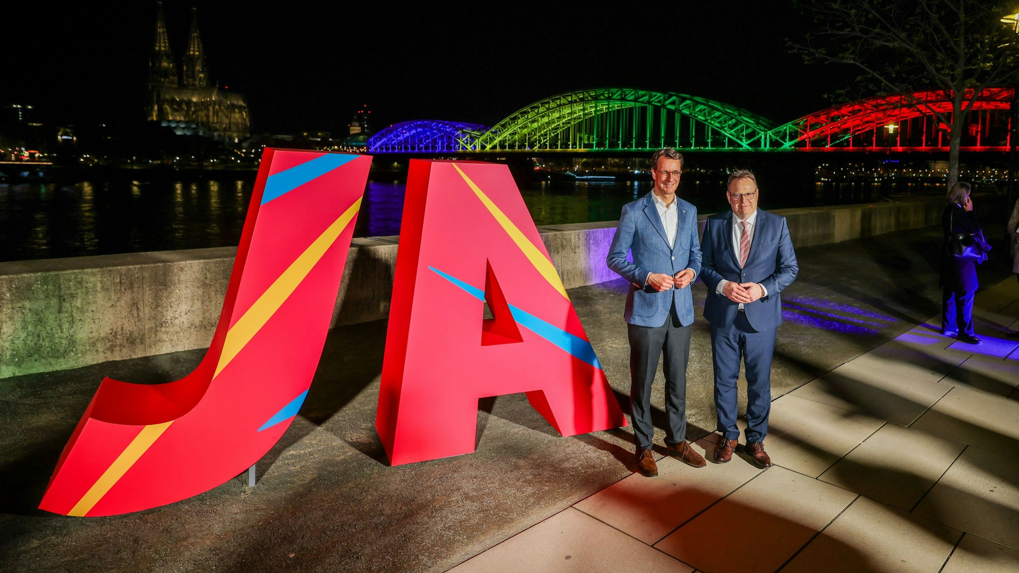 Hendrik Wüst (CDU, l.), Ministerpräsident des Landes Nordrhein-Westfalen, und Torsten Burmester (SPD), Oberbürgermeister der Stadt Köln, stehen vor dem großen „Ja“ und der in den olympischen Farben angeleuchteten Hohenzollernbrücke.