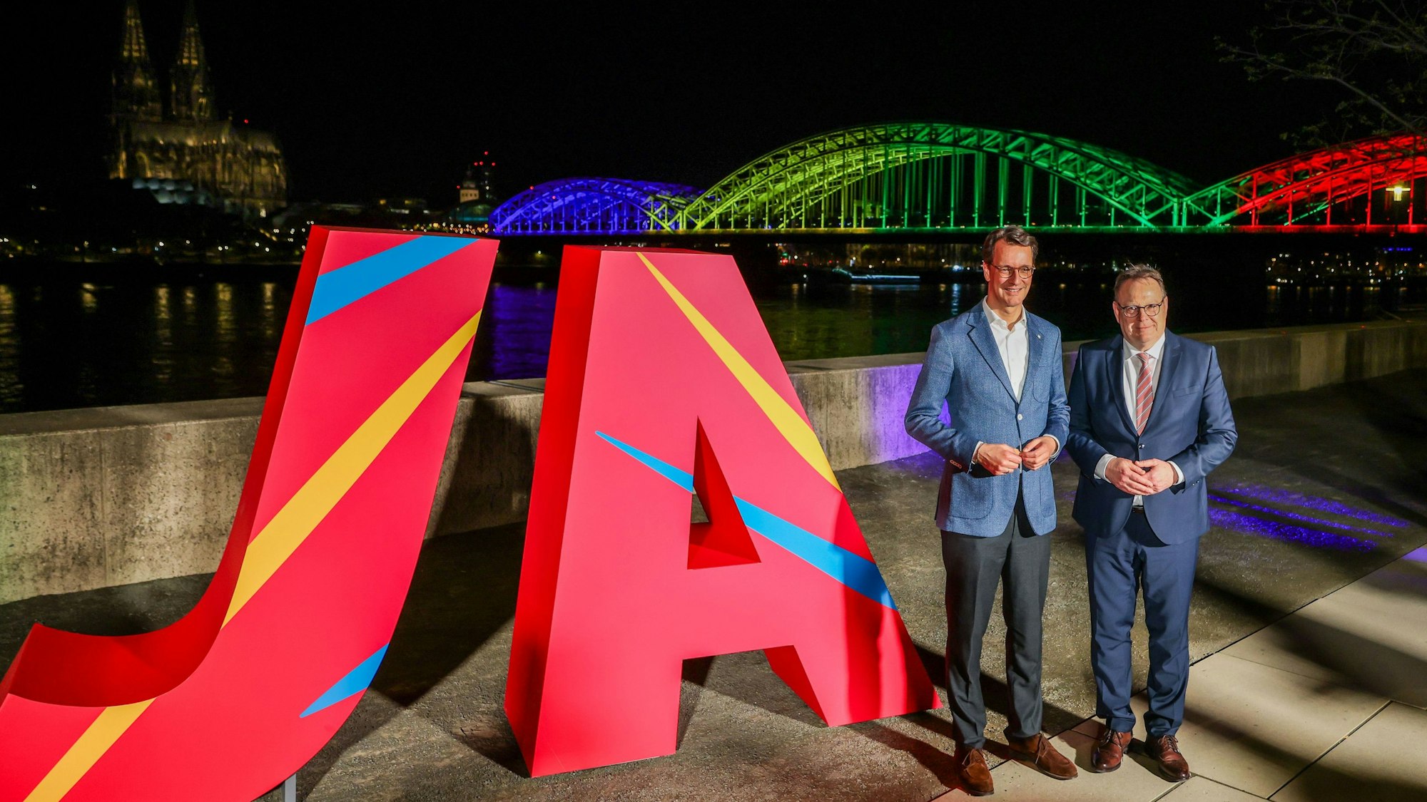 NRW-Ministerpräsident Hendrik Wüst (CDU, links und Torsten Burmester (SPD), Oberbürgermeister der Stadt Köln, stehen vor dem großen „Ja“ und der in den olympischen Farben angeleuchteten Hohenzollernbrücke in Köln.