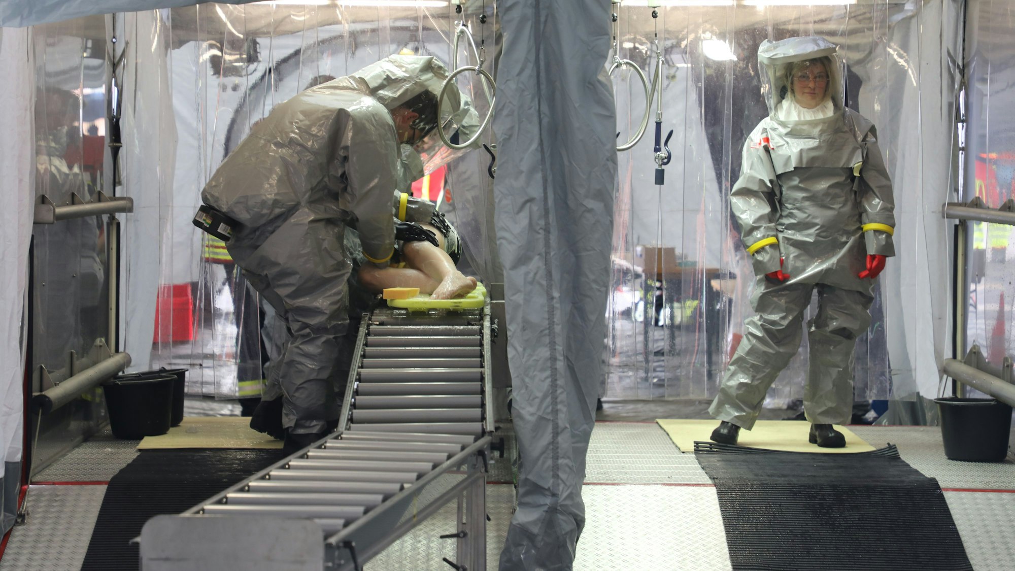 Einsatzkräfte in Schutzkleidung stehen in einem Dekontaminations-Abrollcontainer und duschen kontaminierte Patienten ab.