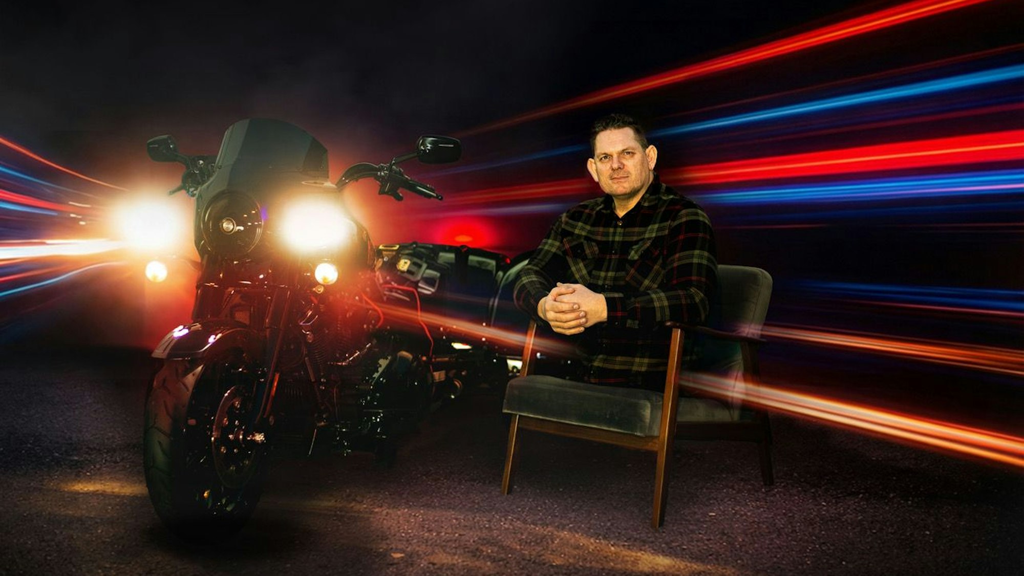 Das Foto zeigt einen Mann, der keine Beine mehr besitzt, auf einem Stuhl. Neben ihm steht ein Motorrad.