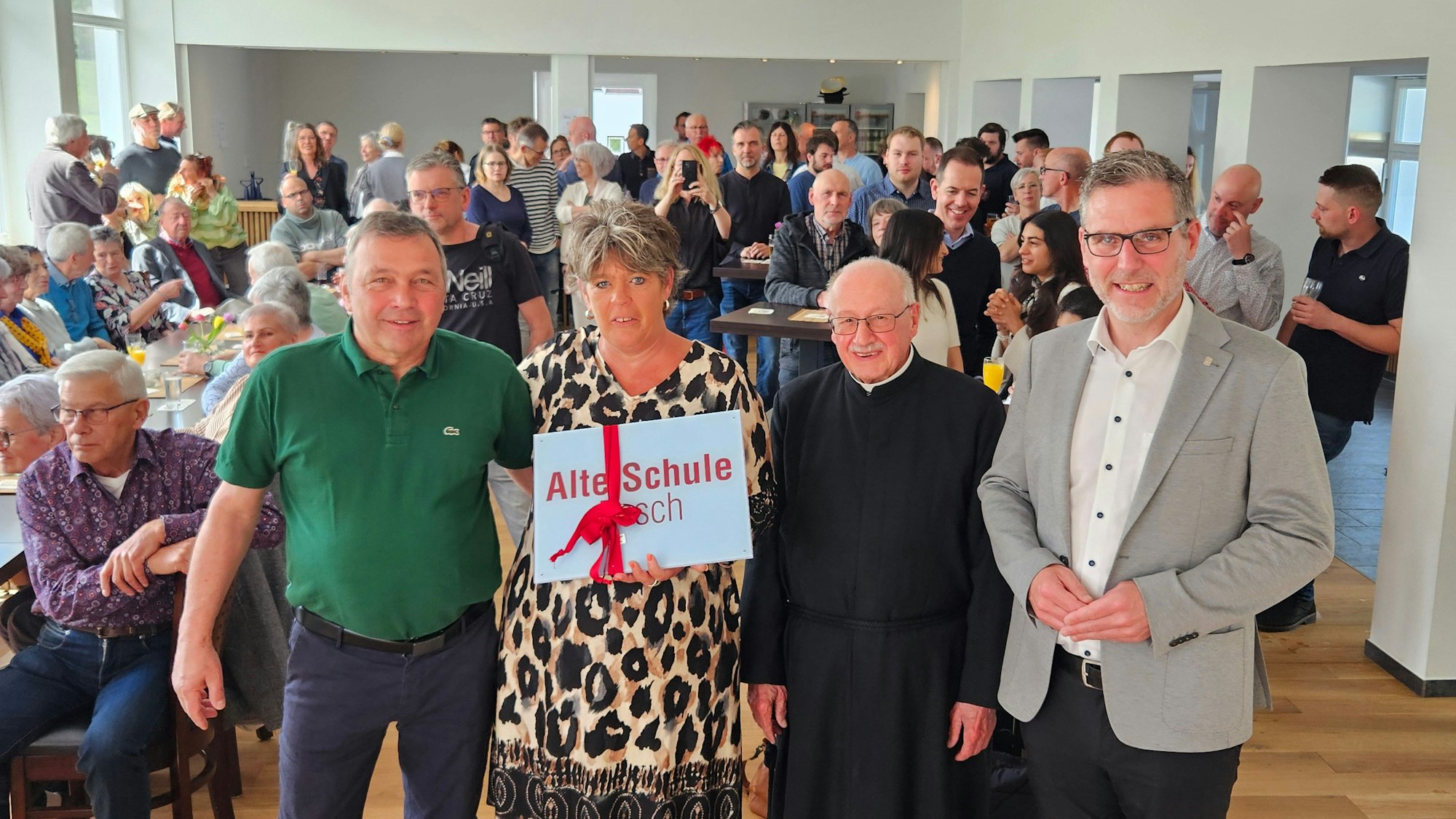 Drei Männer und eine Frau stehen nebeneinander. Die Frau hält ein Schild, auf dem Alte Schule Pesch steht. Im Hintergrund sind die Gäste der Einweihung zu sehen.
