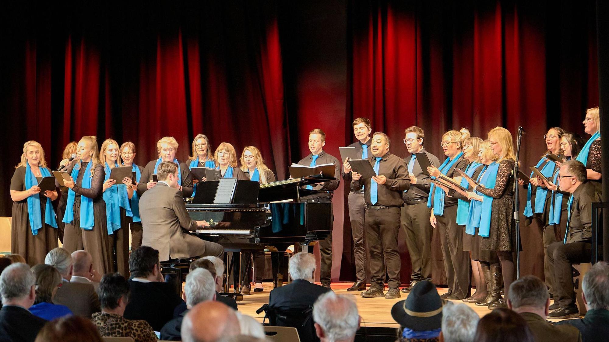 Das Bild zeigt den Chor auf der Bühne mit einem Pianisten.