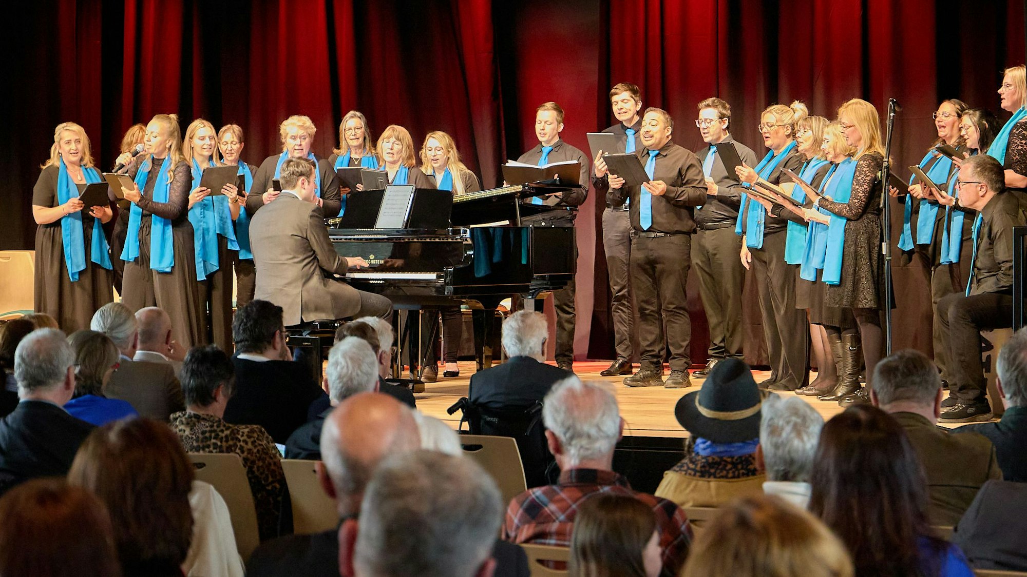 Sängerinnen und Sänger stehen auf der Bühne, vor ihnen sitzt ein Mann am Flügel. Im Vordergrund ist das Publikum von hinten zu sehen.