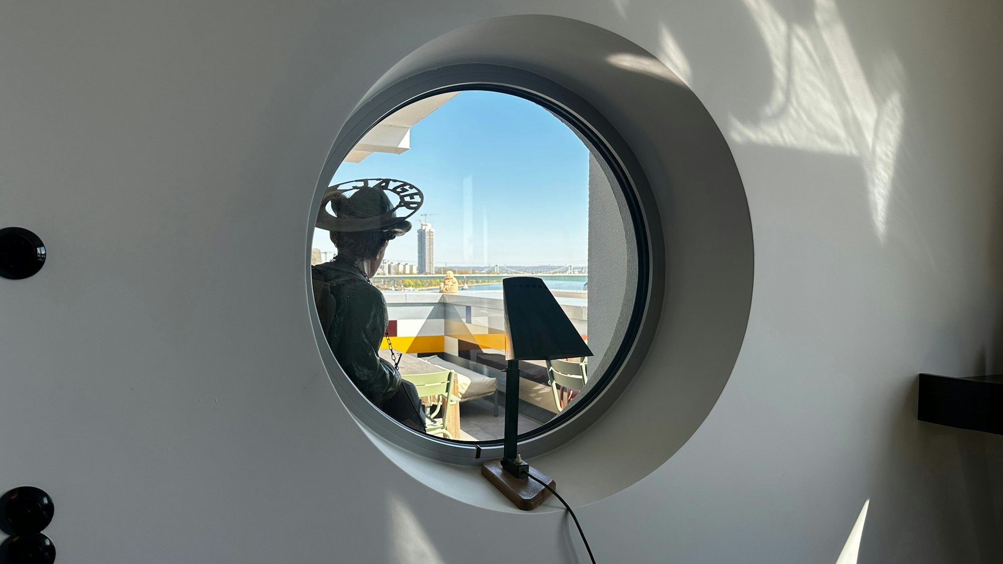 Ein rundes Fenster gibt den Blick frei auf einen Balkon, dahinter der Rhein mit dem Messeturm.