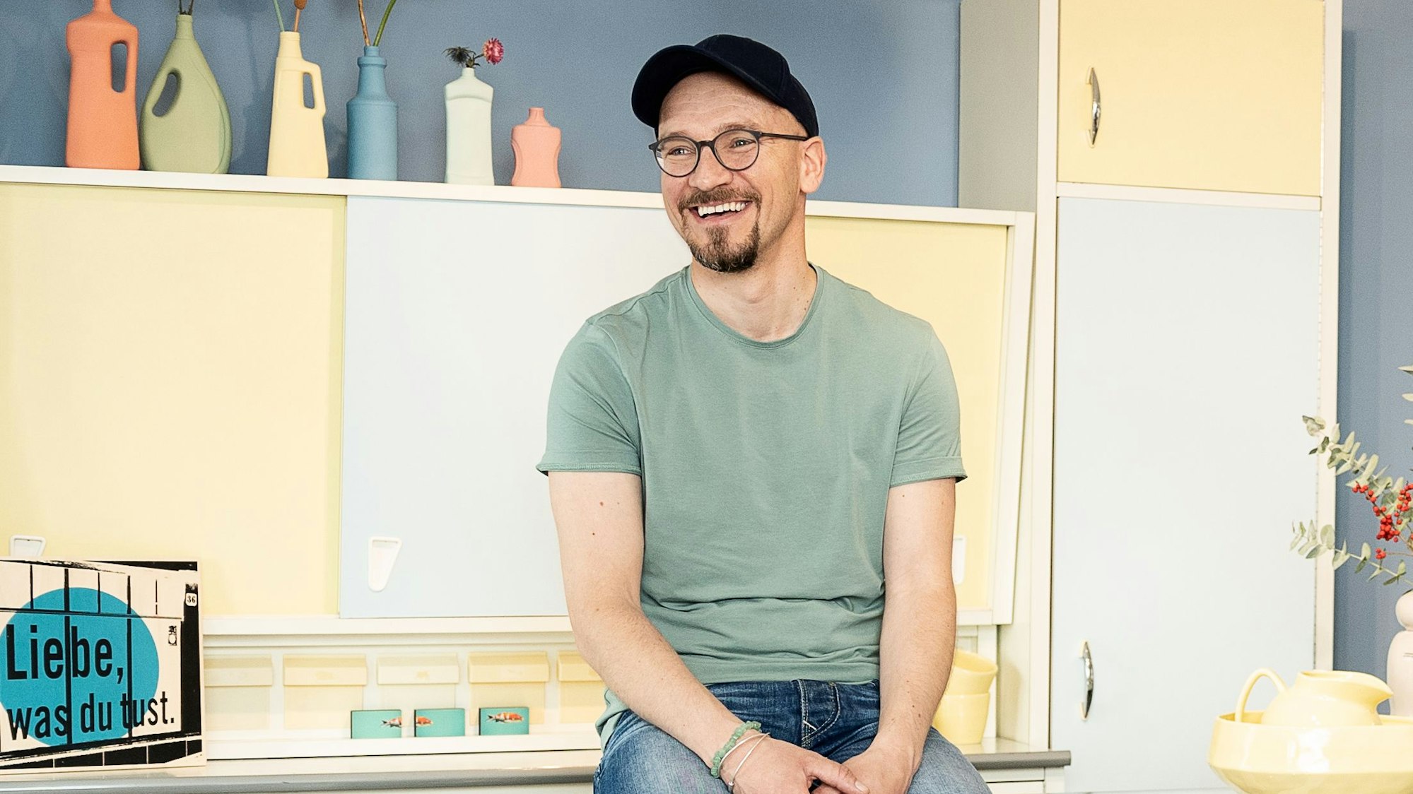 Ein Mann mit blauer Kappe, Brille und Bart sitzt auf einem Küchenbüfett.