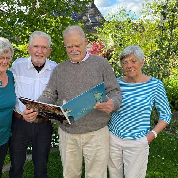 Zwei Frauen und zwei Männer stehen in einem Garten vor grünen Büschen und Bäumen. Einer der Männer hält ein Fotobuch.