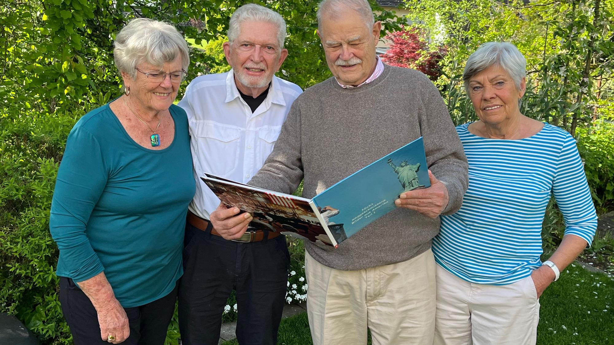 Zwei Frauen und zwei Männer stehen in einem Garten vor grünen Büschen und Bäumen. Einer der Männer hält ein Fotobuch.