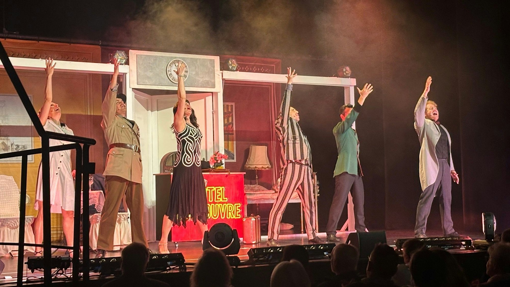 Zu sehen ist eine Theater-Besetzung auf der Bühne. Sie recken jeweils ihren linken Arm in die Höhe in Richtung Scheinwerfer.