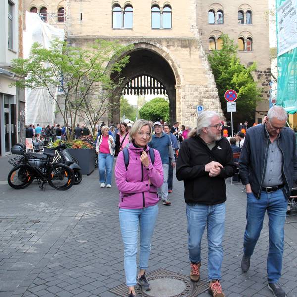 Drei Menschen laufen über den Eigelstein, im Hintergrund ist die Eigelsteintorburg sehen.