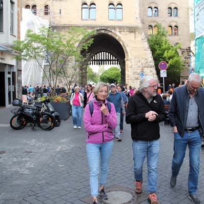 Drei Menschen laufen über den Eigelstein, im Hintergrund ist die Eigelsteintorburg sehen.