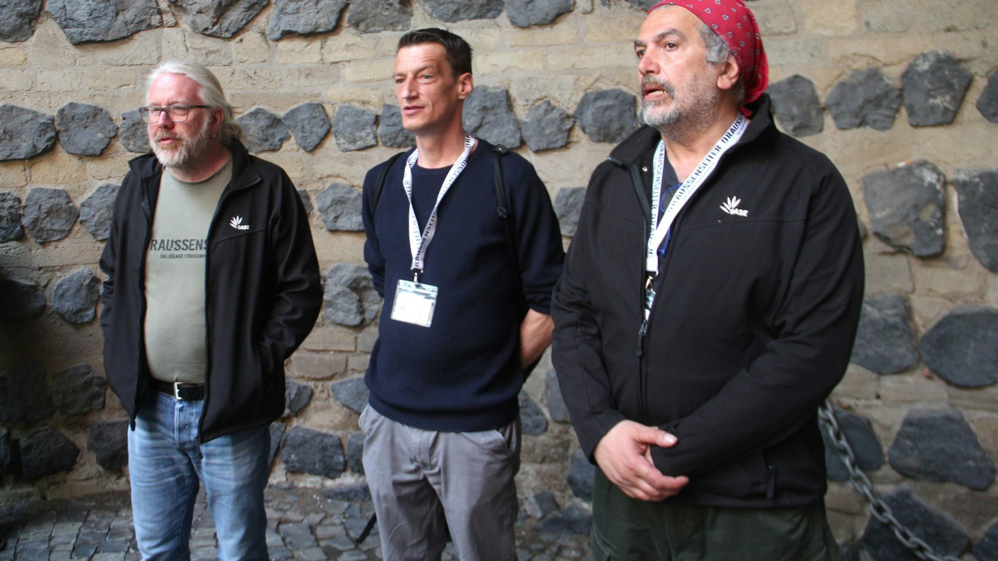 Drei Menschen stehen vor einer Mauer mit groben Steinen.