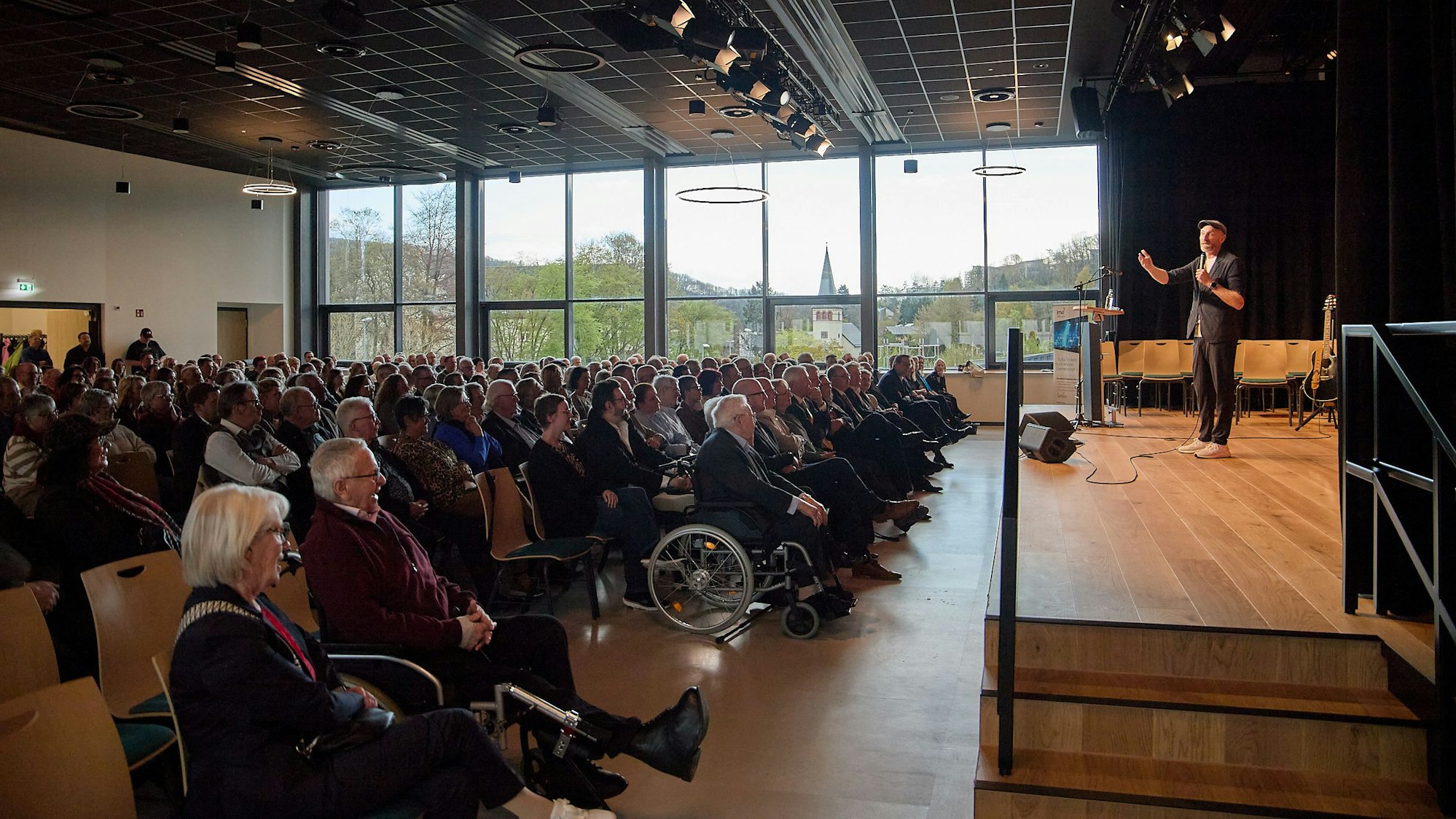 Das Publikum sitzt vor der Bühne, auf der Martin Schopss steht. Durch eine Fensterfront fällt Licht in den Raum.