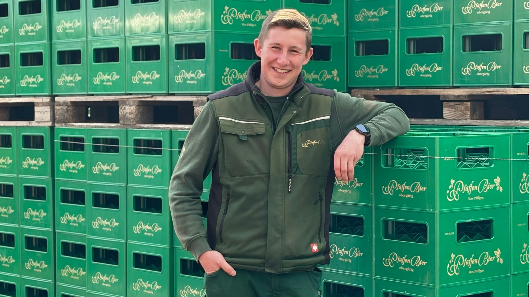 Max Päffgen lehnt mit dem linken Arm an aufgetürmten Bierkästen und lächelt in die Kamera.