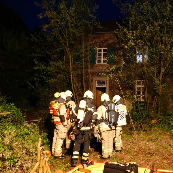 Feuerwehrleute stehen vor dem leerstehenden Haus auf einem zugewucherten Grundstück.