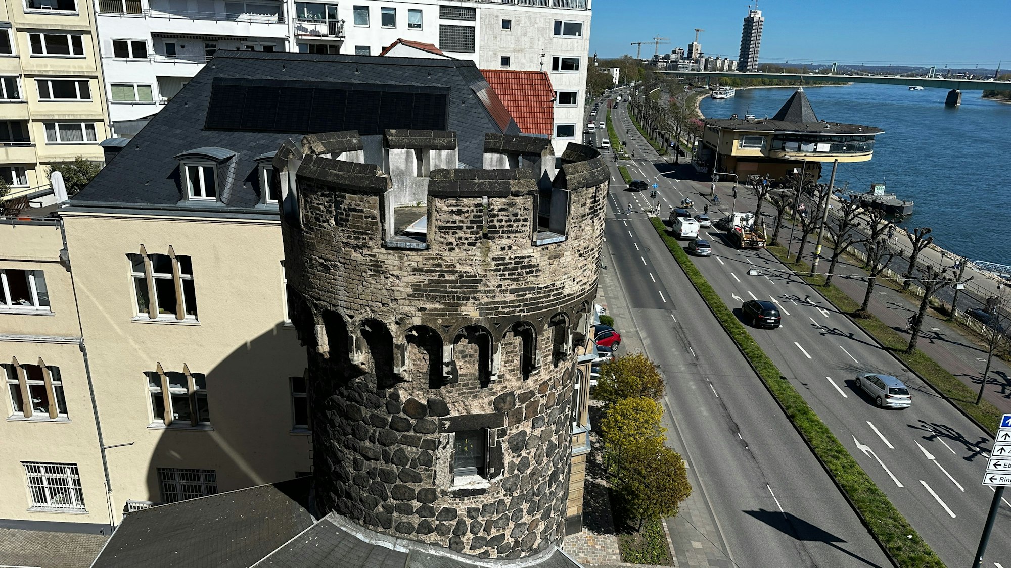 Von einem Balkon aus fällt der Blick auf einen steinernen Turm, eine vierspurige Straße, die Bastei, den Rhein, die Mülheimer Brücke und ein Hochhaus.