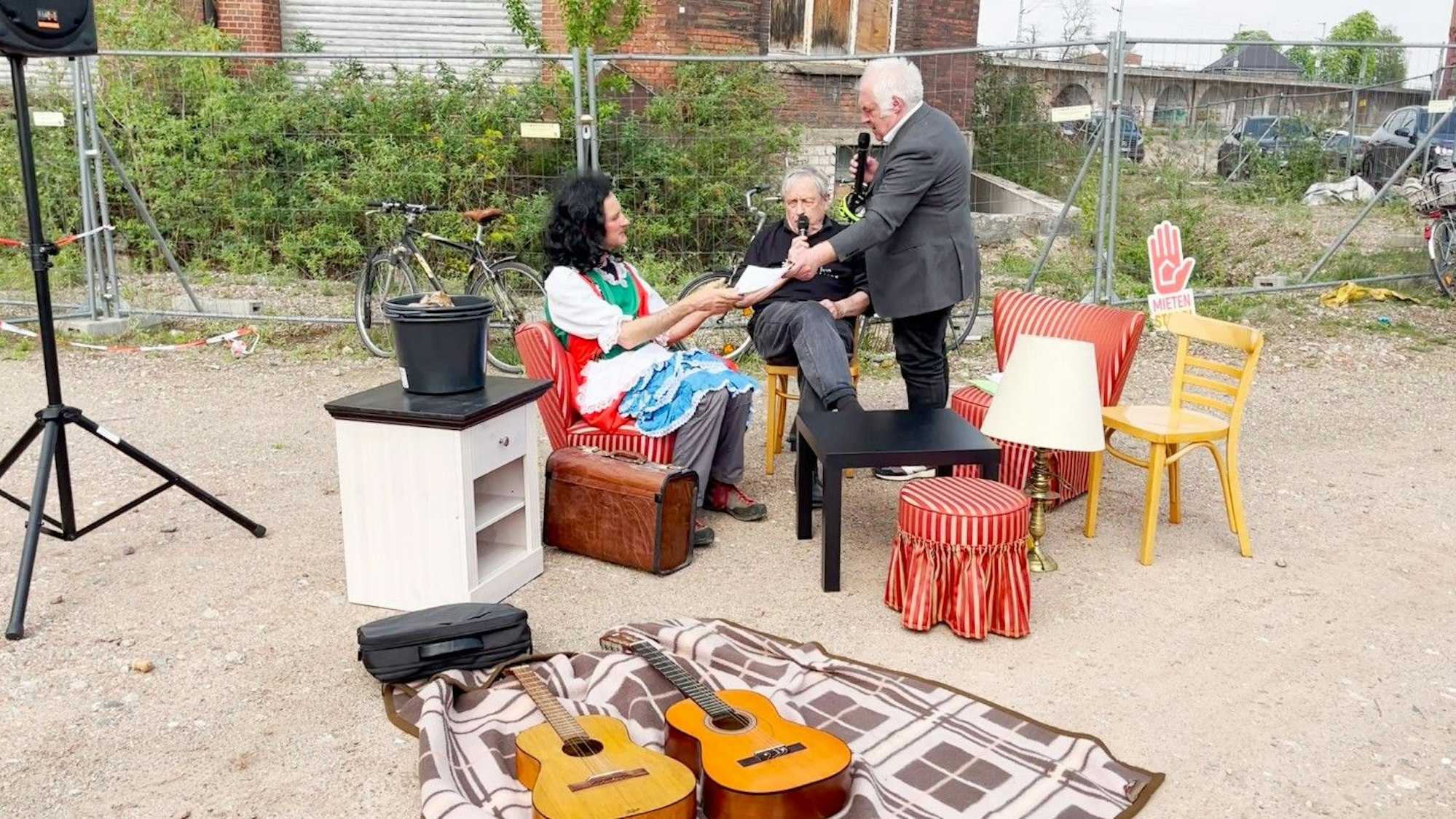 Drei Männer sind in einer Theaterszene zu sehen. Einer mit langer dunkler Perücke bekommt von einem anderen einen Vertrag zur Unterzeichnung überreicht.