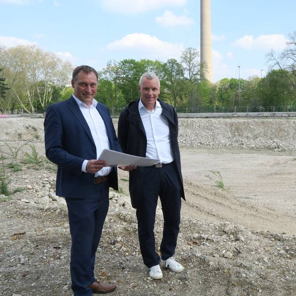 Zwei Männer in Anzügen stehen auf einem leeren Grundstück. Im Hintergrund sind der Colonius und das Telekom-Hochhaus an der Inneren Kanalstraße zu sehen.