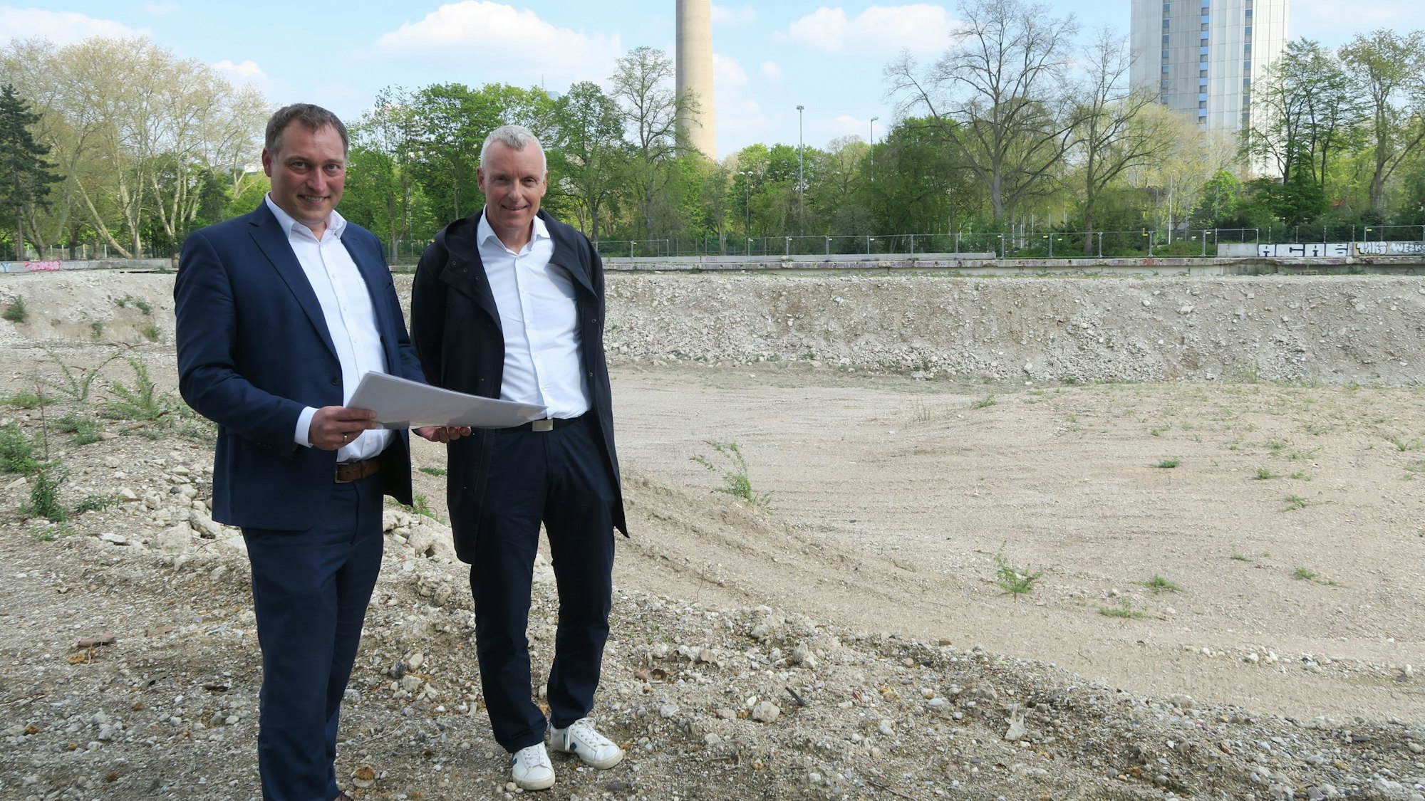 Zwei Männer in Anzügen stehen auf einem leeren Grundstück. Im Hintergrund sind der Colonius und das Telekom-Hochhaus an der Inneren Kanalstraße zu sehen.