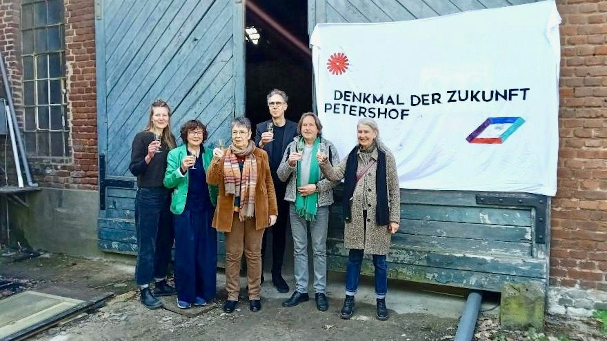 Sechs Menschen stehen mit Sektgläsern in der Hand vor einem alten Gemäuer, an dem ein Transparent hängt: Denkmal der Zukunft, Petershof