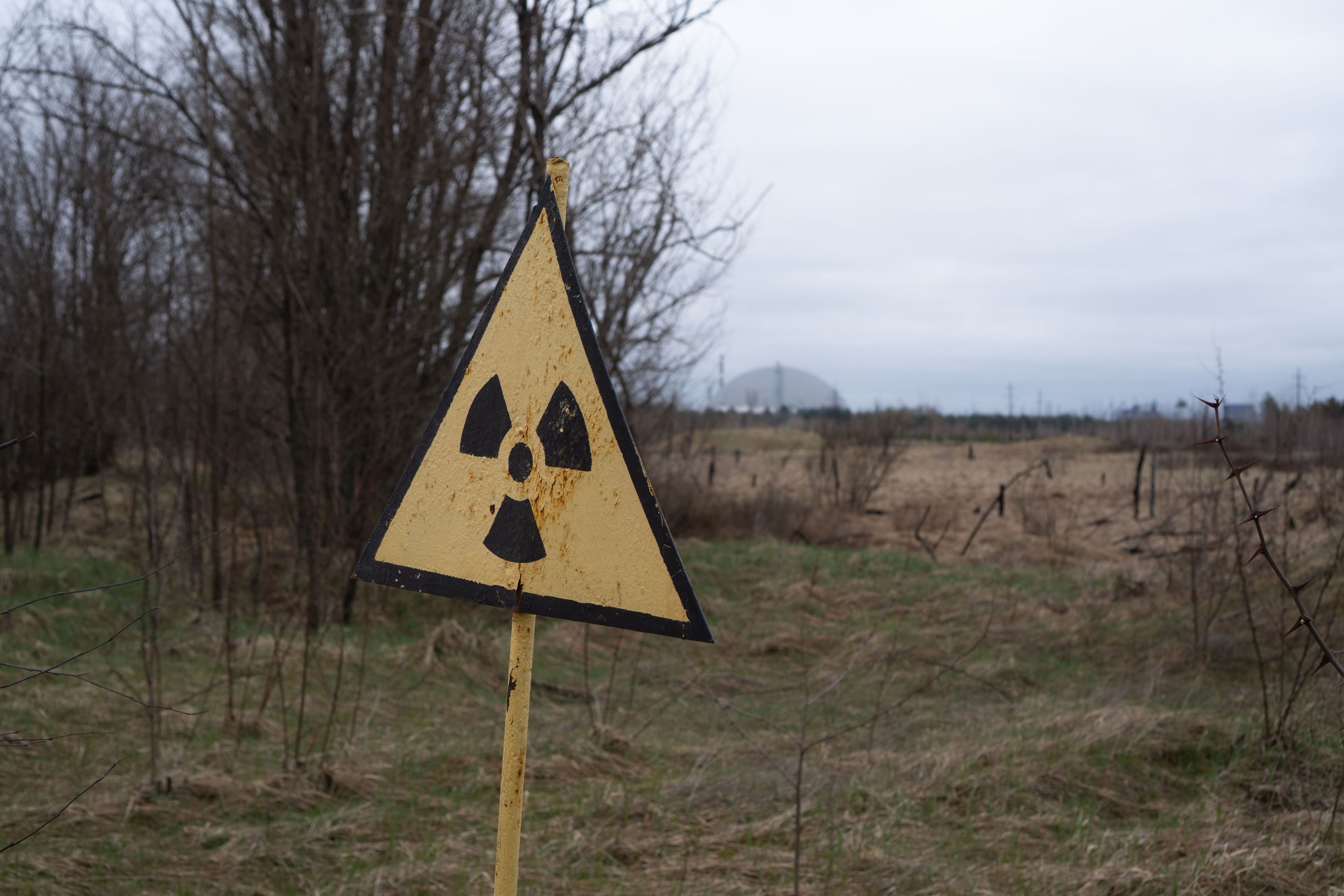 09.04.2026, Ukraine, Tschernobyl: Ein Radioaktivitäts-Warnzeichen, im Hintergrund das Kernkraftwerk Tschernobyl, aufgenommen am 9. April 2026. Was als harmloser Sicherheitstest begann, entwickelte sich am 26. April 1986 zur bedeutendsten Atomkatastrophe der Geschichte. An diesem Tag explodierte Reaktor 4 des Wladimir-Iljitsch-Lenin-Kernkraftwerks, weitläufig bekannt als Kernkraftwerk Tschernobyl. Foto: Nina Mesitz/APA/dpa +++ dpa-Bildfunk +++