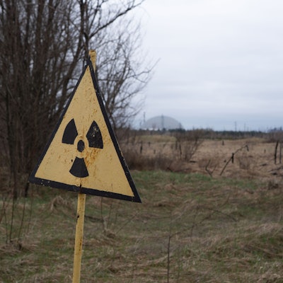 09.04.2026, Ukraine, Tschernobyl: Ein Radioaktivitäts-Warnzeichen, im Hintergrund das Kernkraftwerk Tschernobyl, aufgenommen am 9. April 2026. Was als harmloser Sicherheitstest begann, entwickelte sich am 26. April 1986 zur bedeutendsten Atomkatastrophe der Geschichte. An diesem Tag explodierte Reaktor 4 des Wladimir-Iljitsch-Lenin-Kernkraftwerks, weitläufig bekannt als Kernkraftwerk Tschernobyl. Foto: Nina Mesitz/APA/dpa +++ dpa-Bildfunk +++