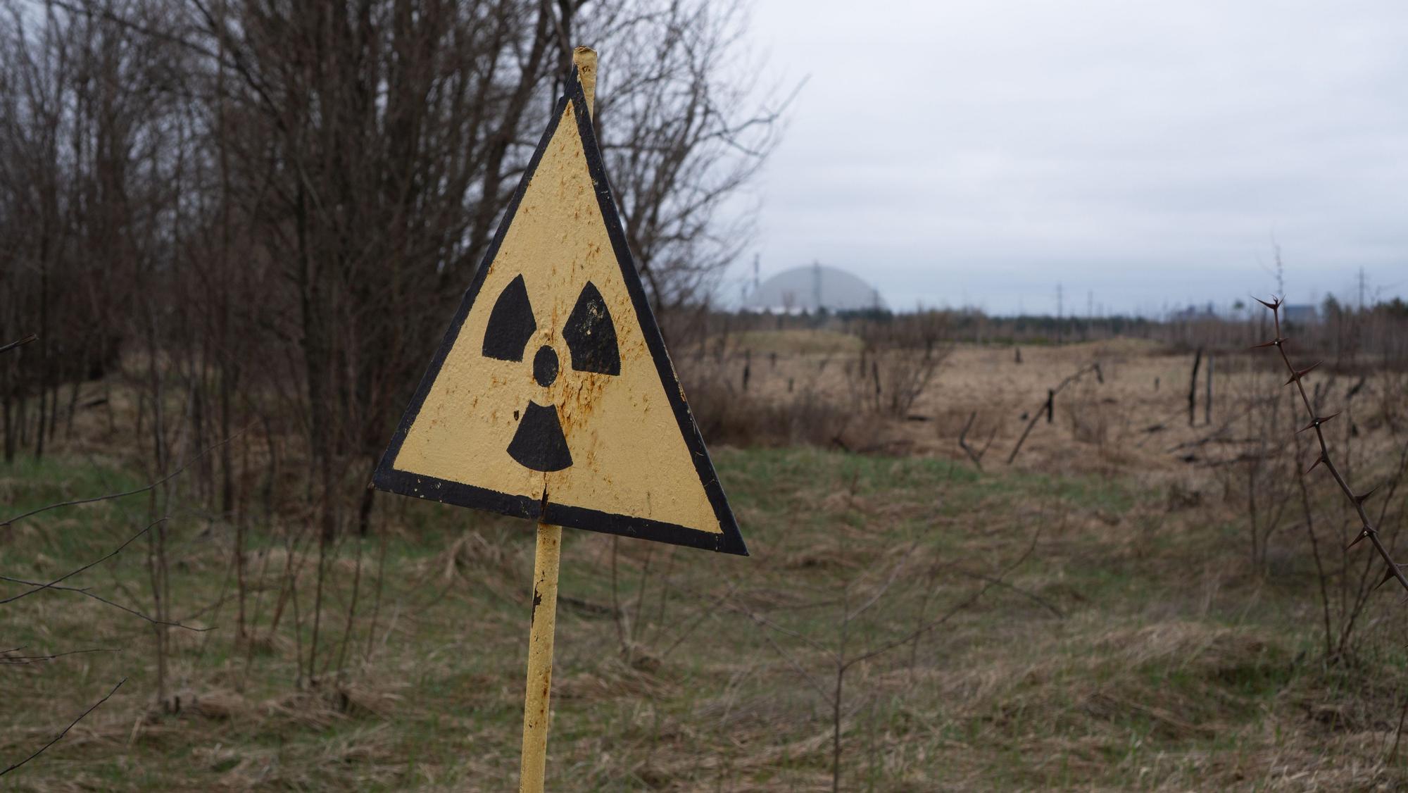 09.04.2026, Ukraine, Tschernobyl: Ein Radioaktivitäts-Warnzeichen, im Hintergrund das Kernkraftwerk Tschernobyl, aufgenommen am 9. April 2026. Was als harmloser Sicherheitstest begann, entwickelte sich am 26. April 1986 zur bedeutendsten Atomkatastrophe der Geschichte. An diesem Tag explodierte Reaktor 4 des Wladimir-Iljitsch-Lenin-Kernkraftwerks, weitläufig bekannt als Kernkraftwerk Tschernobyl. Foto: Nina Mesitz/APA/dpa +++ dpa-Bildfunk +++