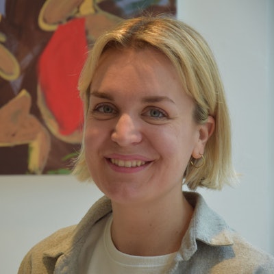 Eine Frau mit blonden, kurzen Haaren steht vor einer weißen Wand, an der ein buntes Gemälde hängt.