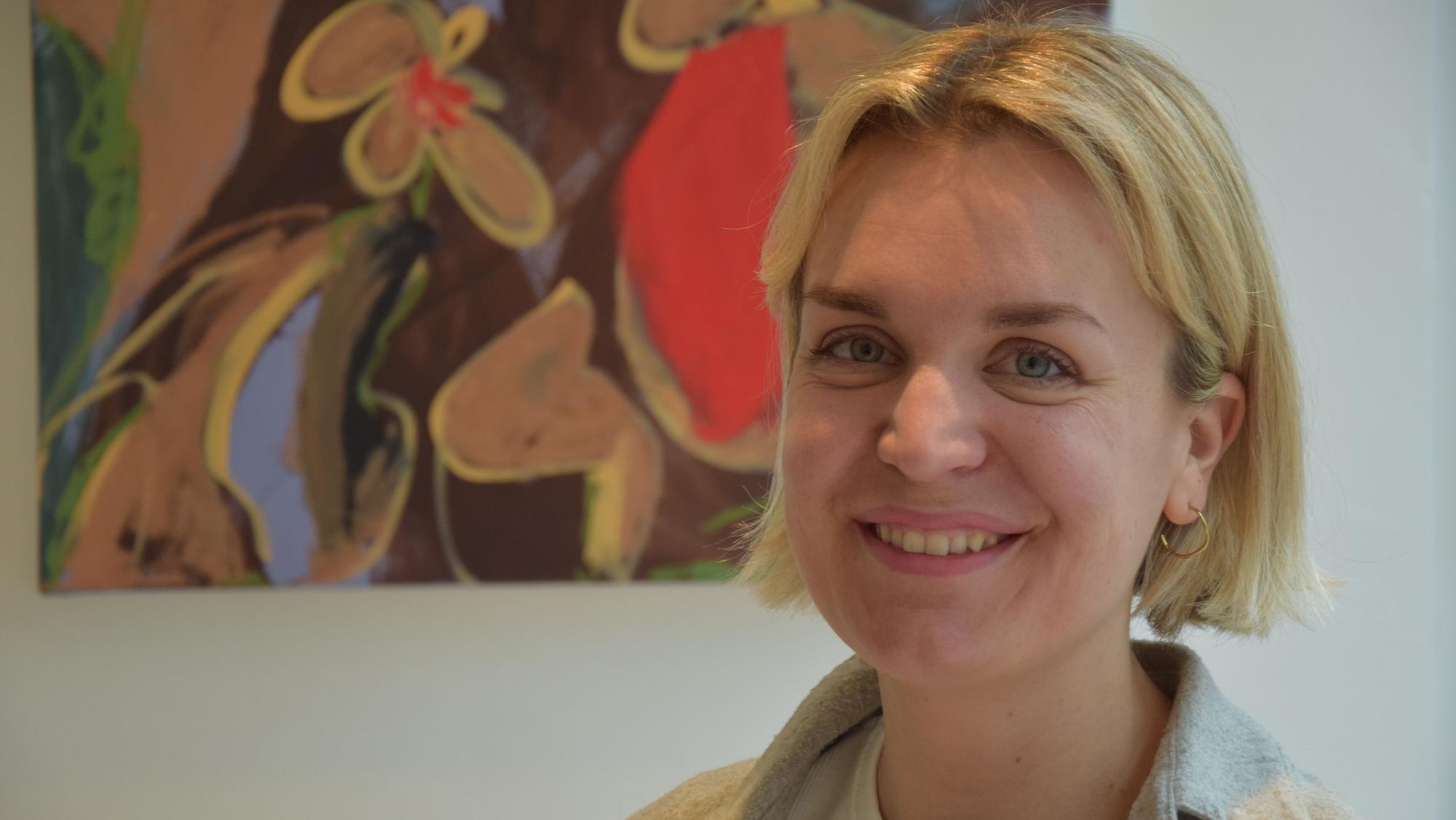 Eine Frau mit blonden, kurzen Haaren steht vor einer weißen Wand, an der ein buntes Gemälde hängt.