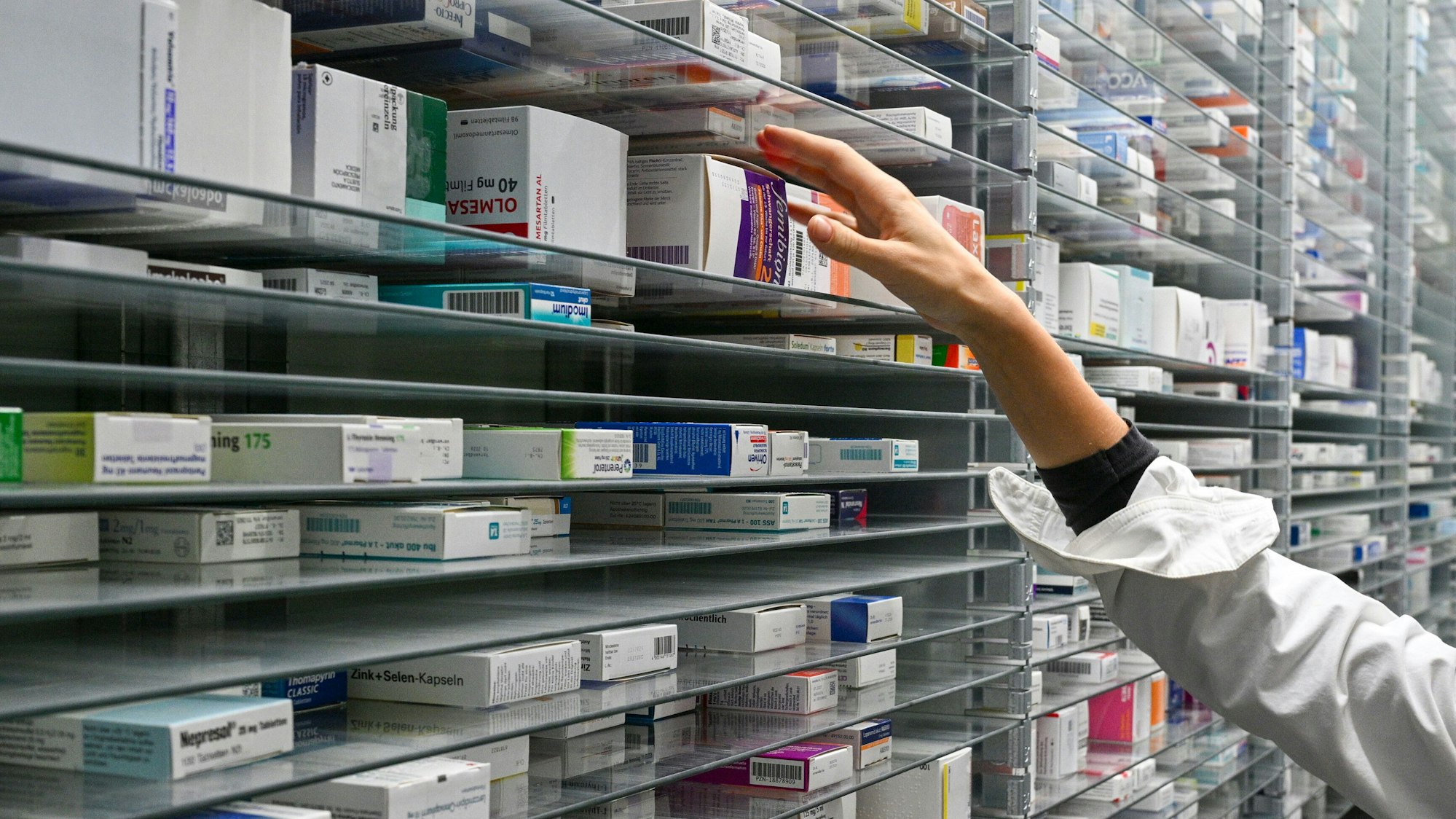 Arzneimittel liegen in einem Medikamentenlager bereit. Eine Hand greift nach einer Packung im Regal.