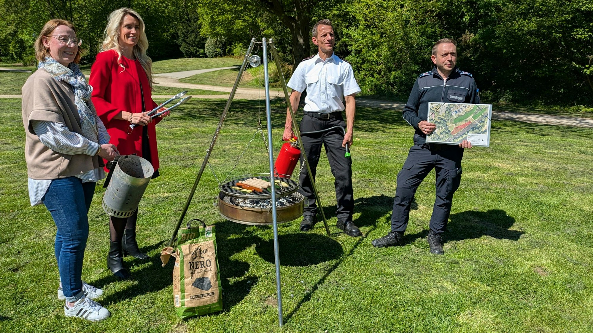 Zwei Frauen und zwei Männer stehen um einen Grill auf einer Wiese.