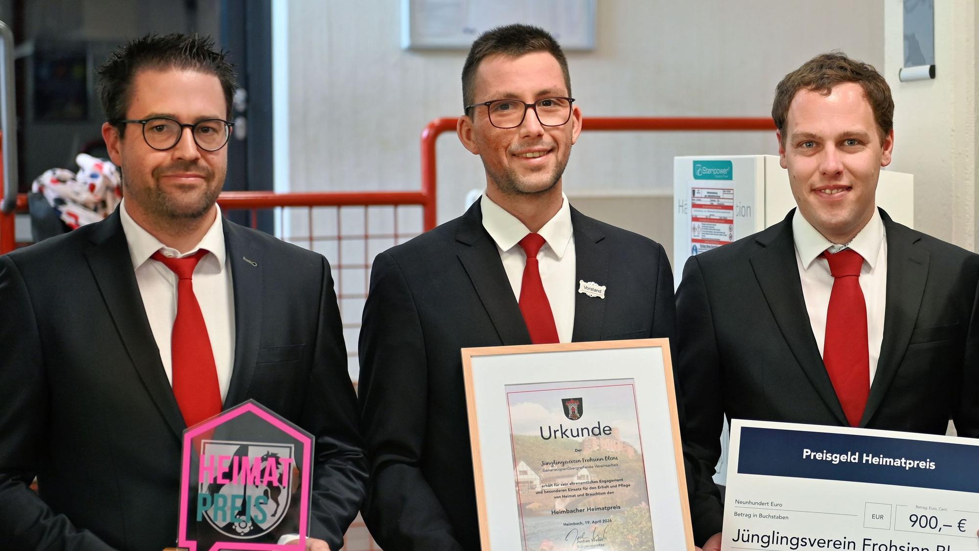 Drei Männer in dunklen Jacketts und roten Krawatten halten den Heimatpreis, die Urkunde und den Scheck in ihren Händen.